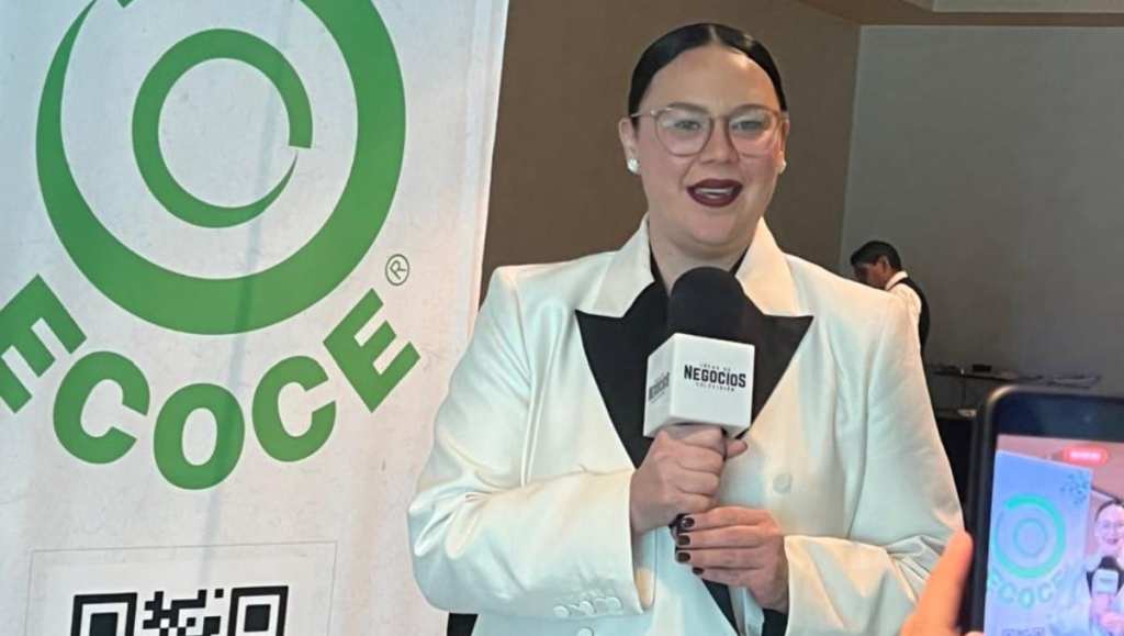 Female reporter in a white blazer and glasses speaks into a microphone labeled 'NEGOCIOS' at a media event, with a large green circular logo beside her.